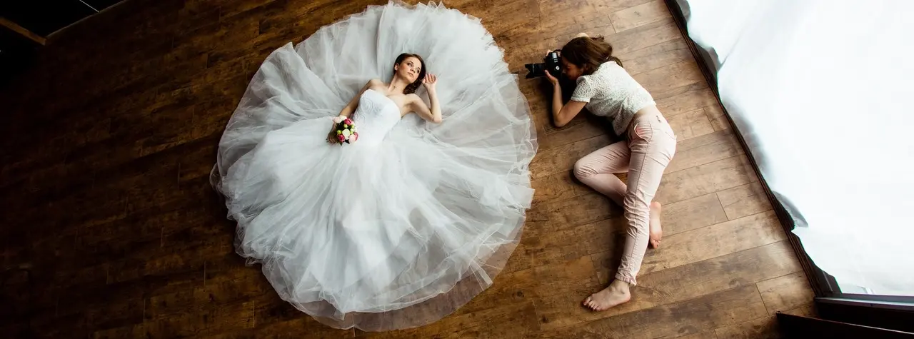 Braut in weißem Kleid liegt auf Holzboden, während Fotografin neben ihr kniet und ein Foto aufnimmt.